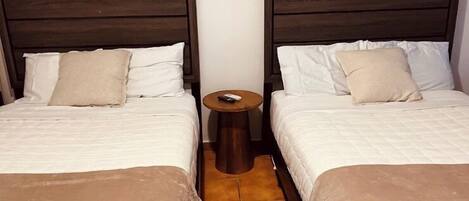 Habitación con dos camas, cada una con cabecero de madera, ropa de cama blanca y una pequeña mesita entre ellas.