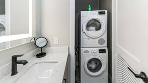 A compact laundry room with two stacked front-loading washing machines, a sink with a modern faucet, and a small round mirror. - Walk to Bengals, Reds, Free Parking, King Bed, Gym (Cincinnati)