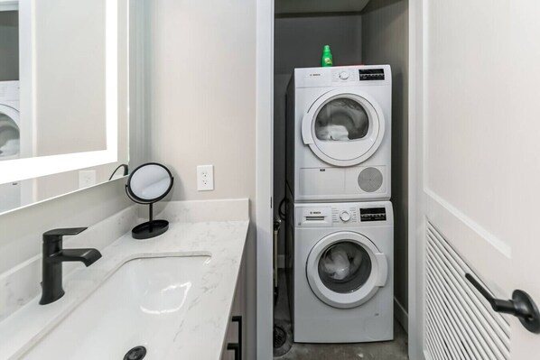 A compact laundry room with two stacked front-loading washing machines, a sink with a modern faucet, and a small round mirror.