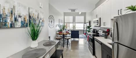 Una cocina moderna con electrodomésticos de acero inoxidable, un comedor con una mesa redonda y sillas, y una sala de estar con un ventanal grande.