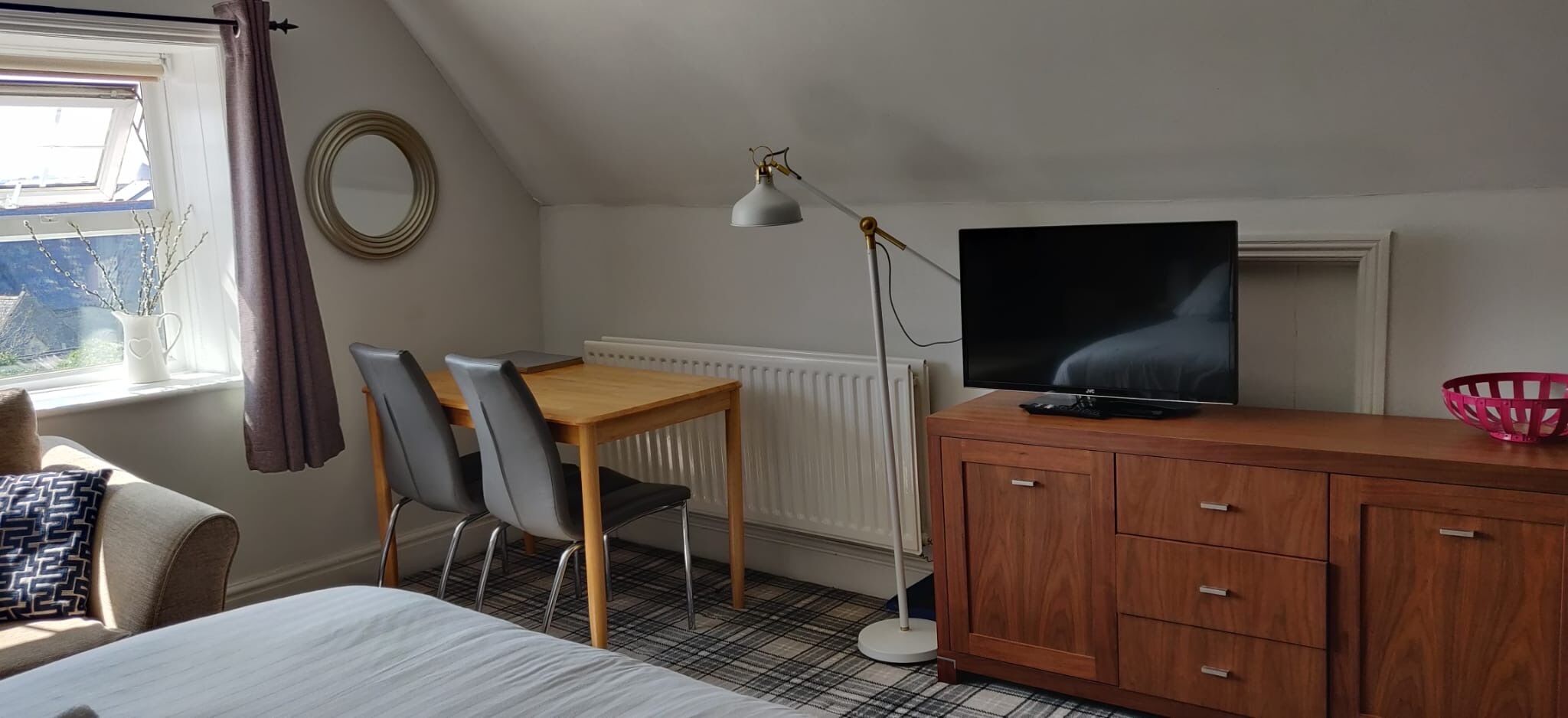 Et værelse med seng, skrivebord med to stole, fjernsyn, radiator og et vindue med gardiner.