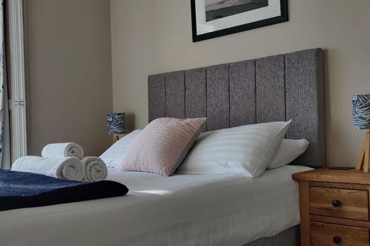 A neatly made bed with pillows, a wooden nightstand with a lamp, and a blue blanket.