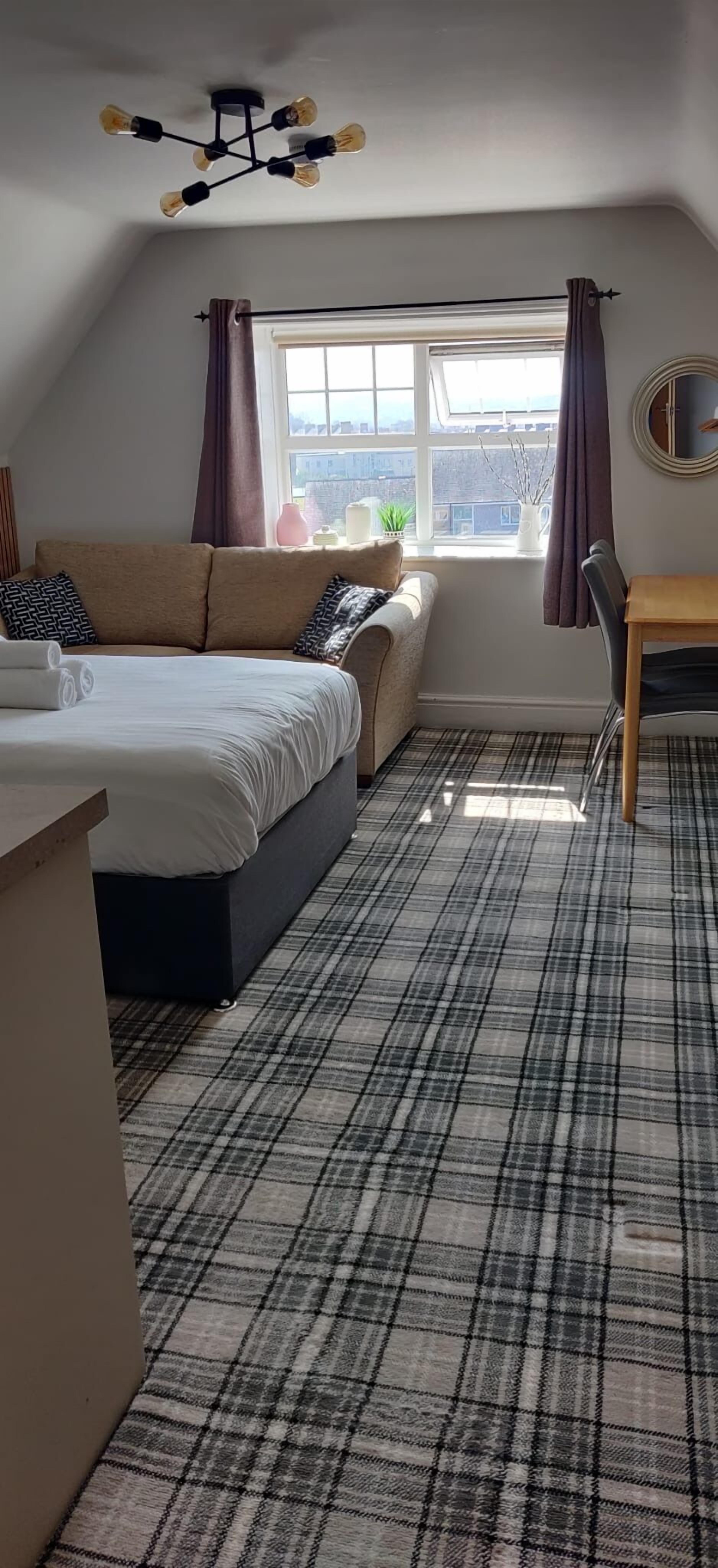 A bedroom with a bed, a desk, a chair, a window with curtains, and a view of the city.