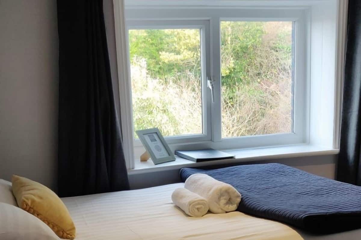 A neatly made bed with a white comforter and a folded towel, a bedside table with a lamp, and a window with curtains.