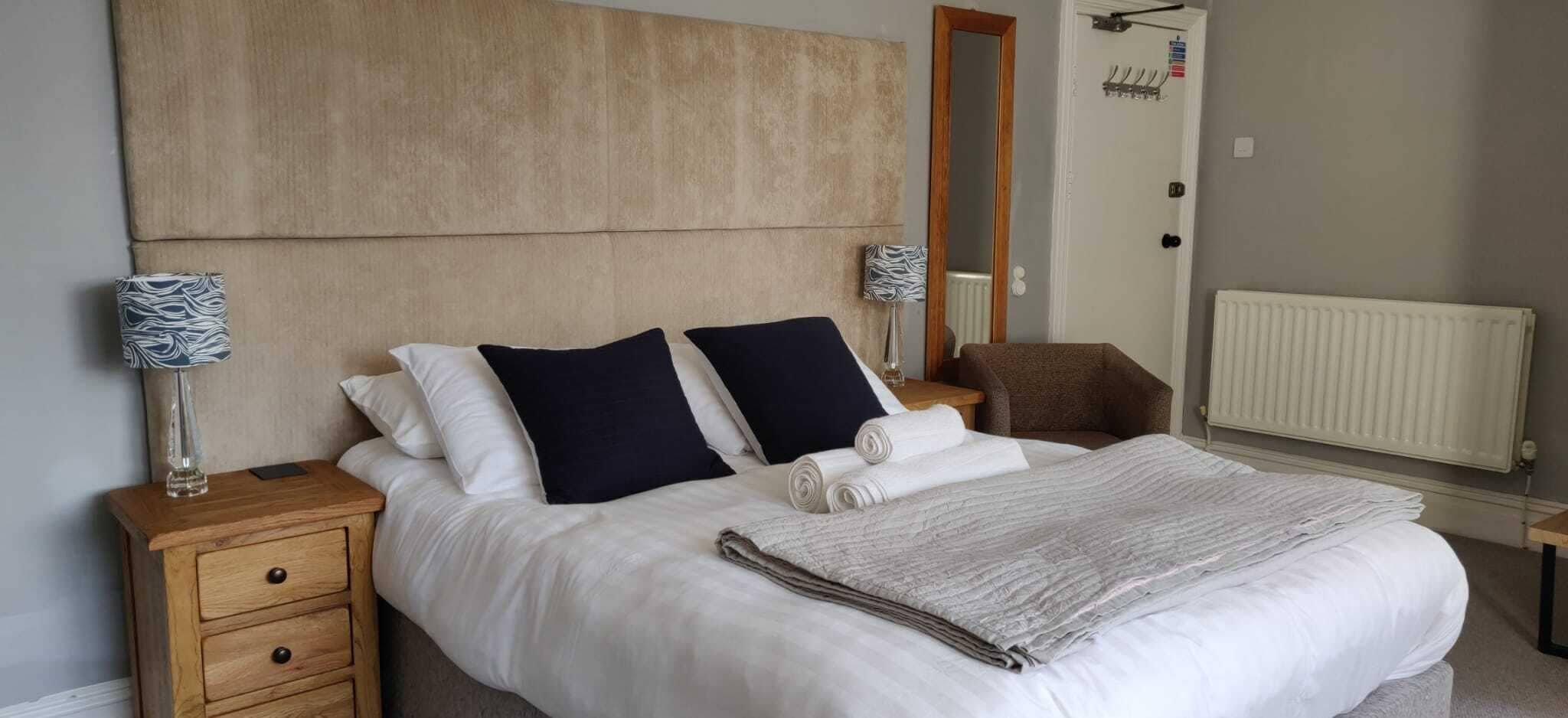 Un lit bien fait, avec des oreillers blancs et bleu marine, une table de chevet en bois avec une lampe, et une chaise marron.