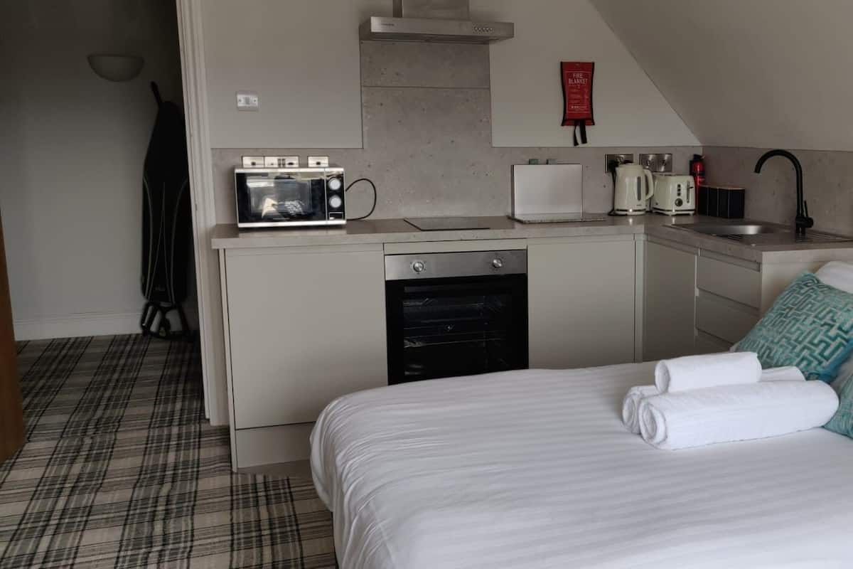 A compact hotel room with a kitchenette, a bed with white linens, and a plaid-patterned floor.