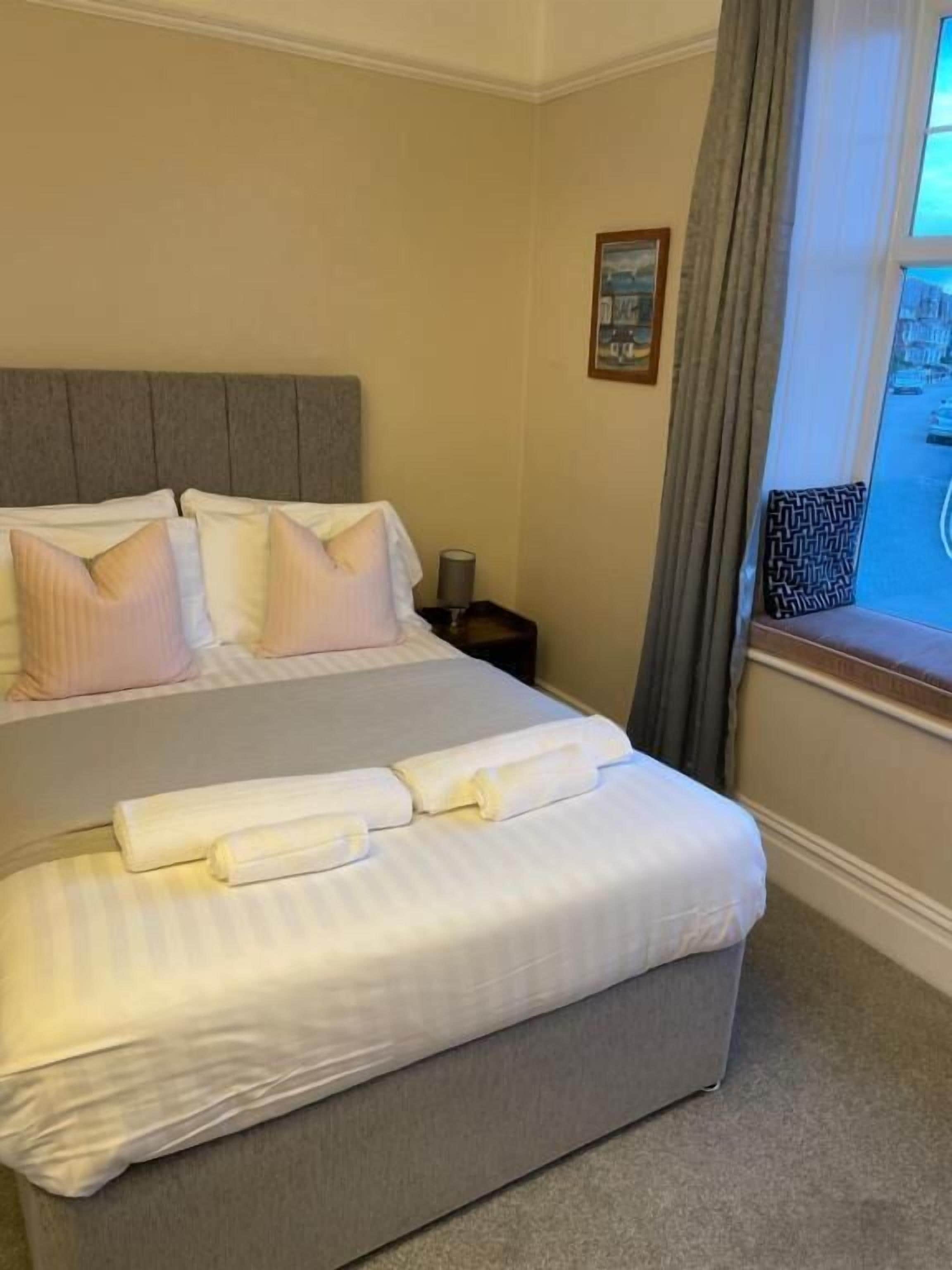Una cama bien hecha con almohadas y una toalla enrollada, una mesita de noche con una lámpara y una ventana con cortinas.