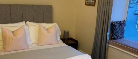 Una cama bien hecha con almohadas y una toalla enrollada, una mesita de noche con una lámpara y una ventana con cortinas.