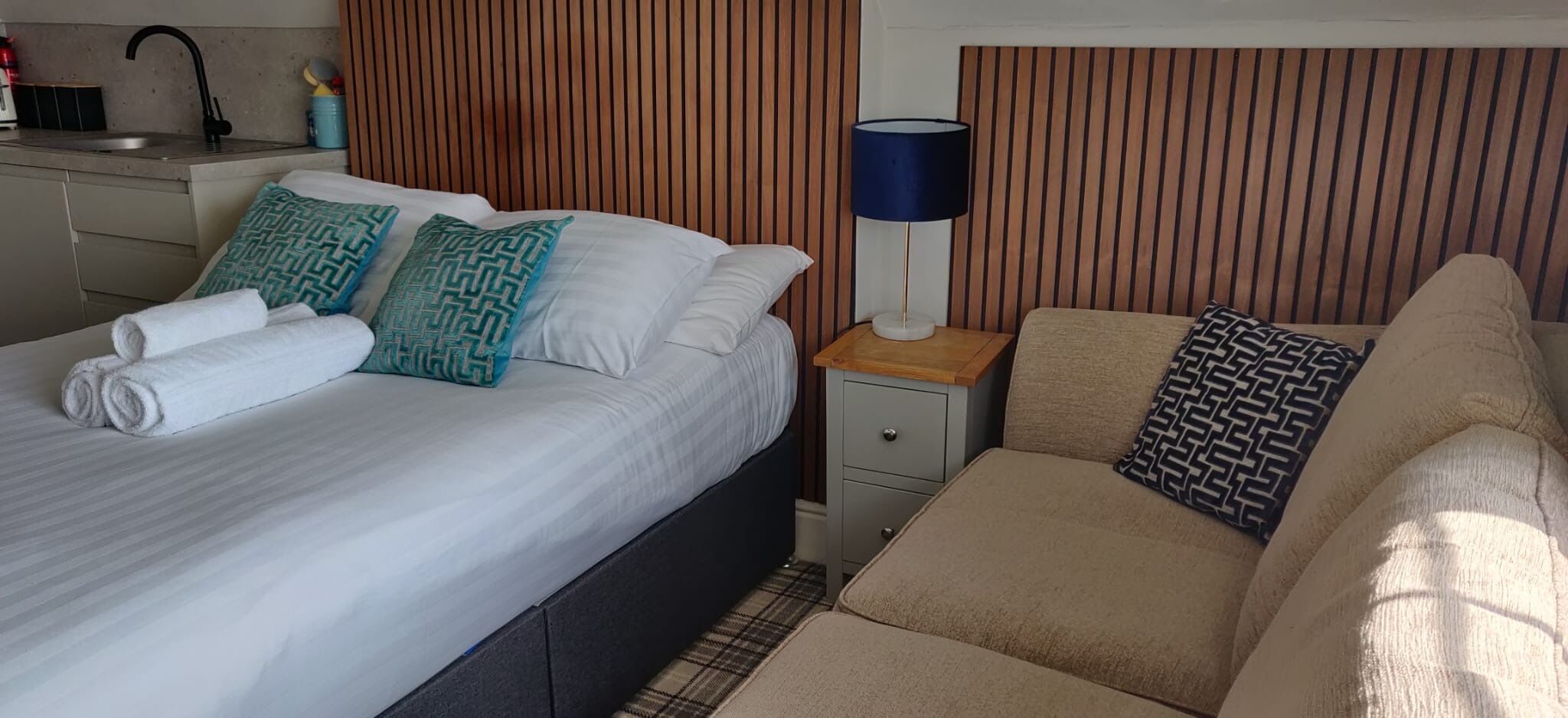 A hotel room with a bed, a bedside table with a lamp, a sofa, and a small table with a lamp.