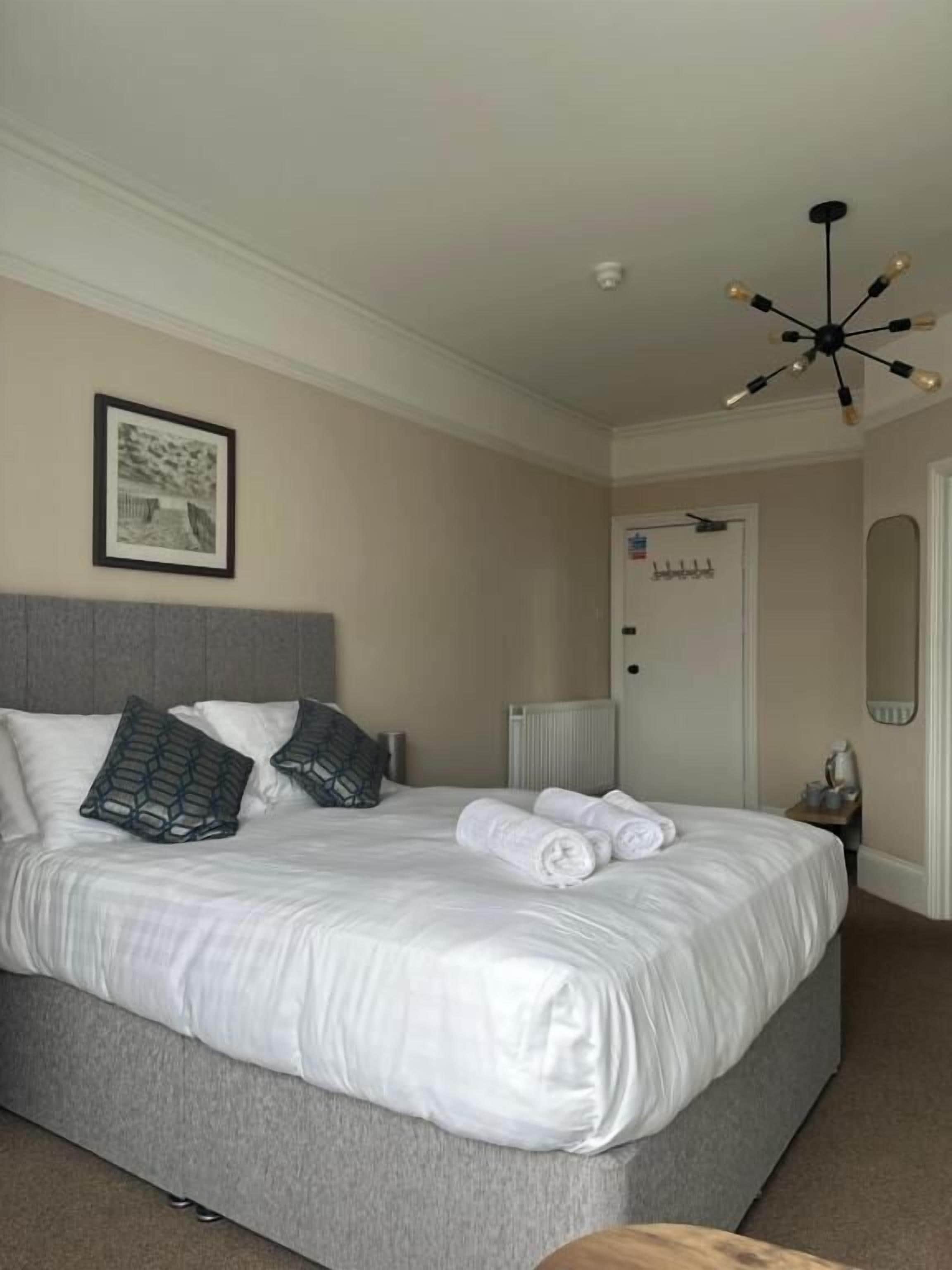 A neatly made bed with white linens and two decorative pillows, a modern ceiling light fixture, and a framed picture on the wall.