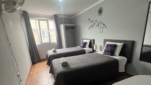 Een hotelkamer met twee bedden, een kleine tafel en een stoel. Er is een raam met gordijnen, een plafondventilator en een wanddecoratie.