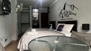 Hotelkamer met een bed, een rond glazen tafeltje, een televisie, een plafondventilator en een muur met een berg afbeelding.