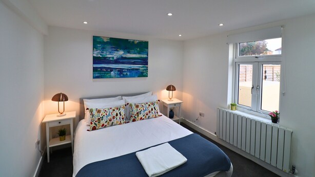 A bedroom with a bed, two bedside lamps, a window, a radiator, and a painting on the wall. - London Gorgeous Two Bedroom Apartment (Mitcham Junction)