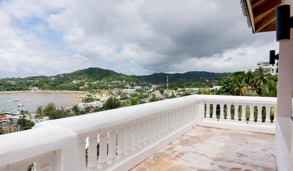 A balcony with a white balustrade overlooking a coastal town with buildings, a hill, and a body of water.