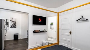 Ein modernes Hotelzimmer mit einem Bett, einem an der Wand montierten Fernseher, einer kleinen Ablage für Toilettenartikel und einer Tür mit Kleiderhaken.