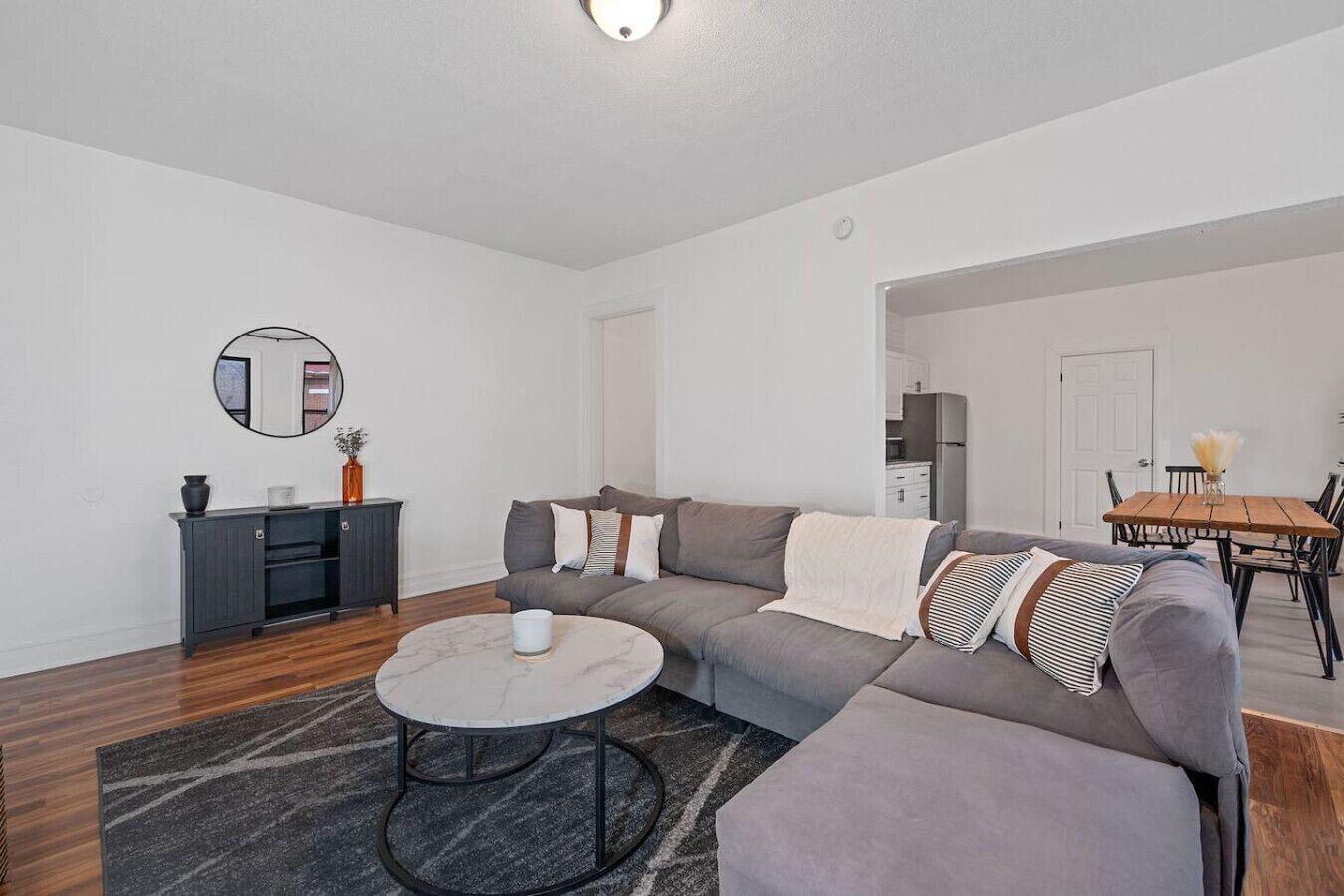 A modern living room with a sectional sofa, a round coffee table, a dark console table, and a dining area with a wooden table and chairs.