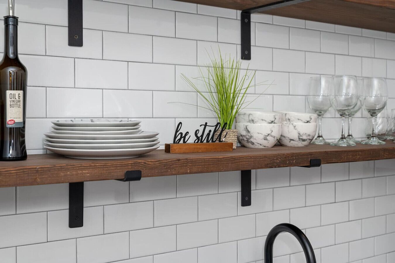 En køkkenhylde med en olieflaske, stablede tallerkener, en plante og et skilt med teksten 'be still'.