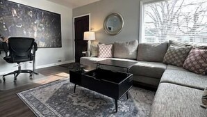 A modern living room with a grey sofa, a black coffee table, and a black office chair. There is a large window with blinds, a round mirror on the wall, and a painting on the wall. - 2 King Beds | Near Sugar House & Downtown (South Salt Lake)