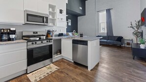 Una cocina moderna con armarios blancos, microondas integrado, estufa y lavavajillas. Hay una pequeña isla con un lavamanos y una cafetera. La cocina está conectada a una sala de estar con un sofá gris y una planta.