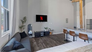 Una habitación de hotel moderna con un sofá gris, un televisor montado en la pared, una mesita con un espejo y una zona de kitchenette con taburetes de bar.
