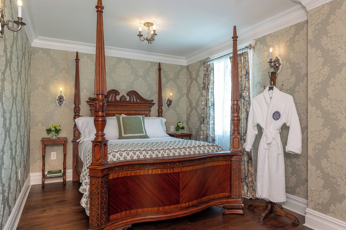 A four-poster bed with a wooden frame and a green and white patterned bedspread. There is a small bedside table with a book and a vase of flowers. A white bathrobe hangs on a stand to the right of the bed.