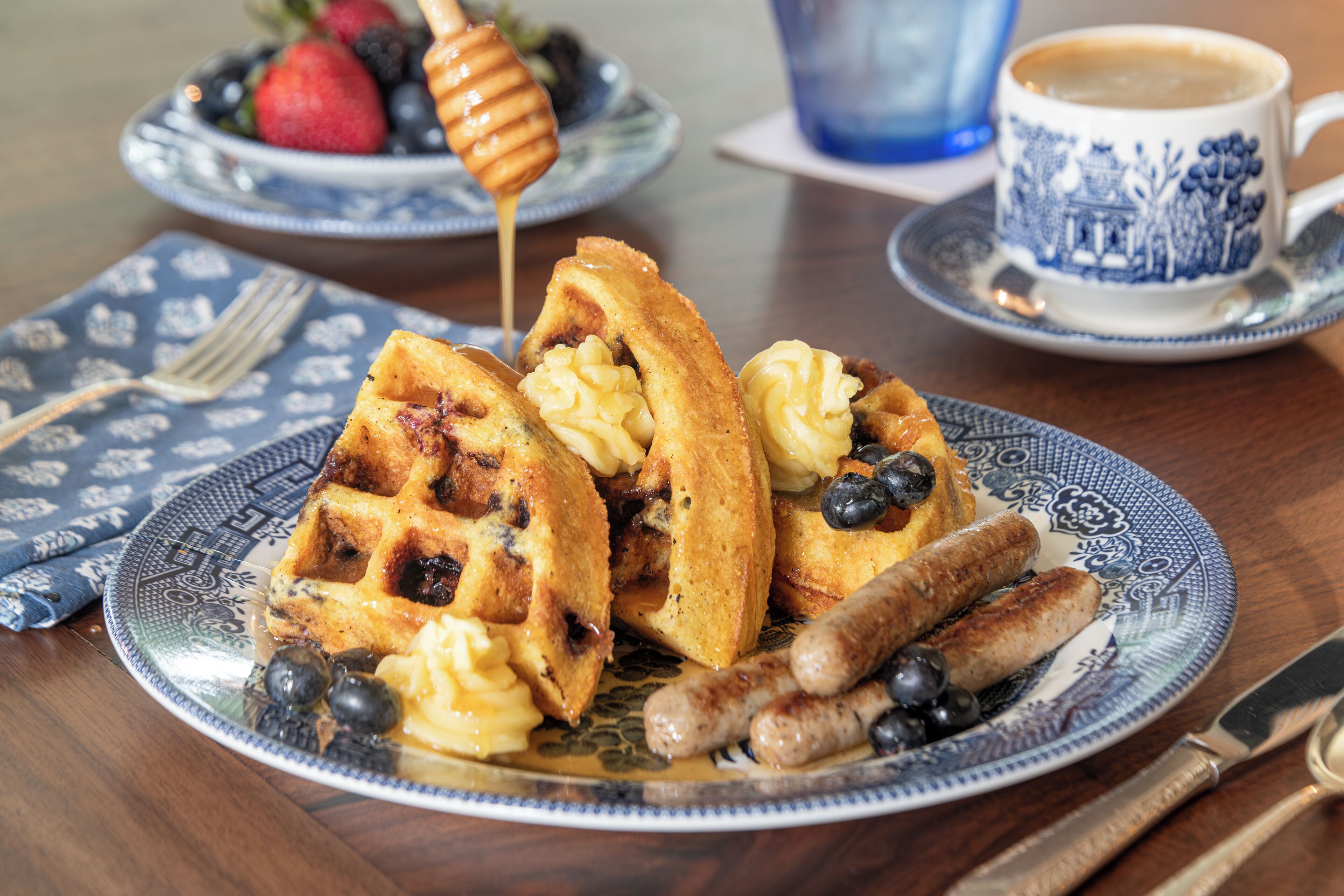 A plate of waffles with syrup and blueberries, a cup of coffee, and sausages on a table.