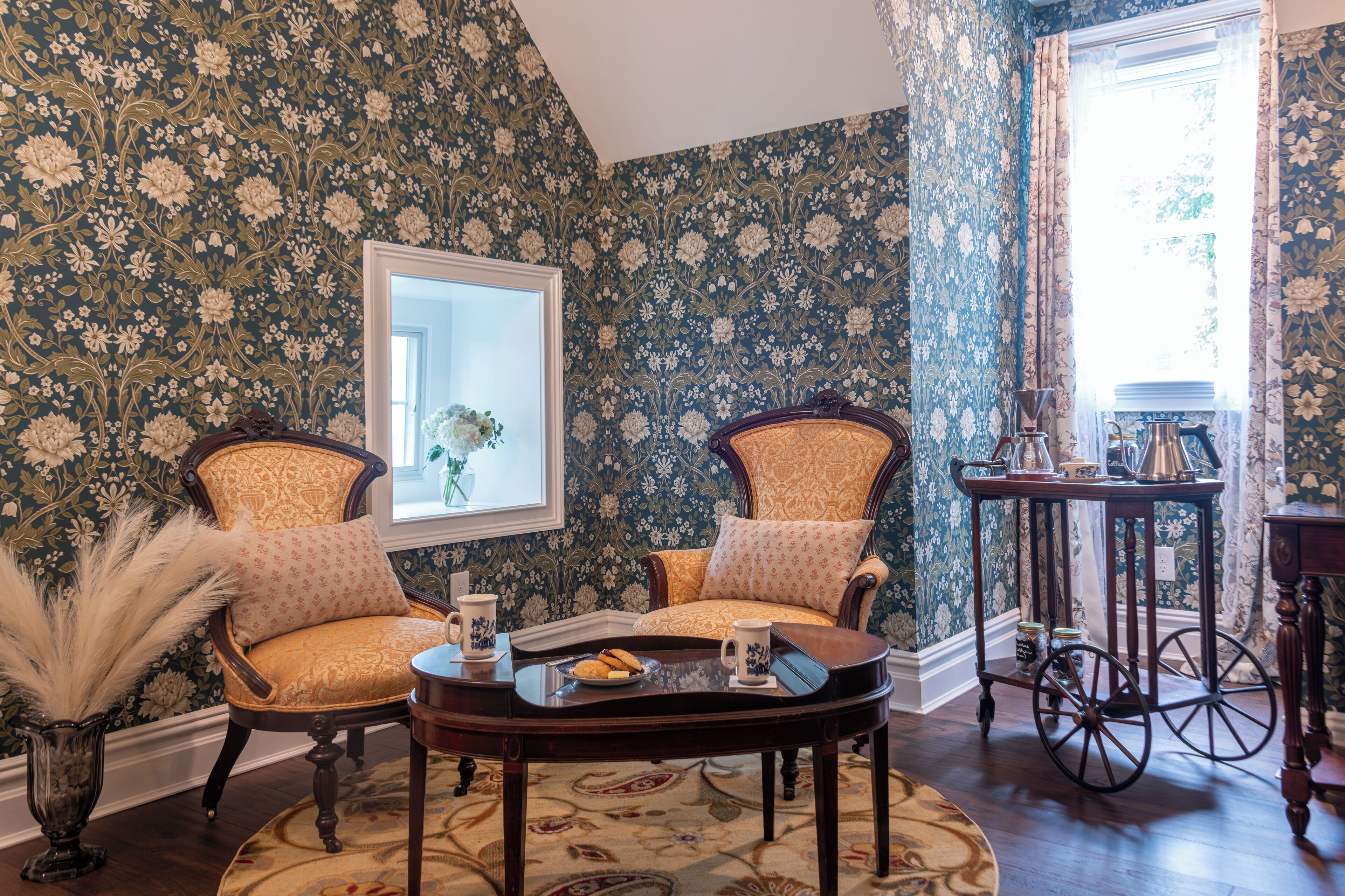 A room with floral wallpaper, two upholstered chairs, a small round table with a tray, a vase with pampas grass, and a window with curtains.