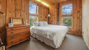 Un dormitorio de cabaña de madera con cama, cómoda y dos ventanas.