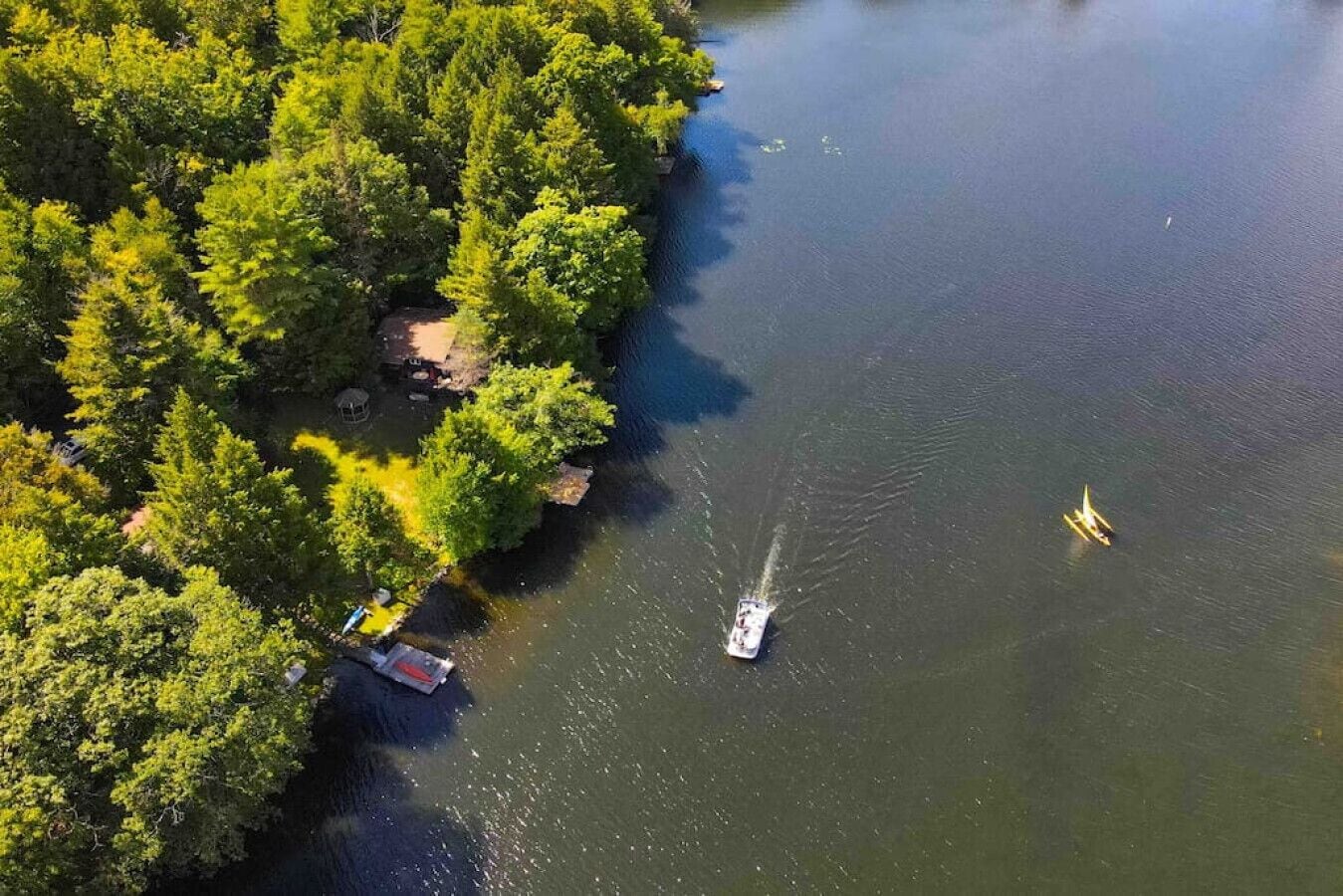 An aerial view of a lake surrounded by dense forest. A boat is visible on the water, and there are two small boats anchored near the shore.
