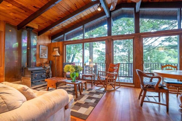 A cozy cabin interior with a wooden ceiling, large windows, a dining area, and a fireplace. - Forest Hollow | Waterfront • Kayaks • Wood Stove (Monterey)