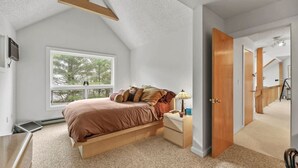 A bedroom with a bed, bedside table, lamp, and a window with a view of trees.