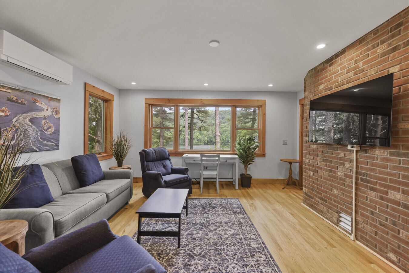 A modern living room with a sofa, armchairs, a coffee table, and a desk. There are large windows with a view of trees, a brick wall with a mounted TV, and a ceiling with recessed lighting.