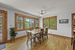 A dining area with a wooden table and chairs, large windows with a forest view, and a ceiling fan. - Bidwell Landing | Lakefront • Spacious (Monterey)