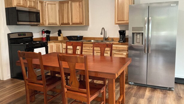 A modern kitchen with wooden cabinets, a stainless steel refrigerator, a microwave, and a stove. There is a dining table with chairs.
