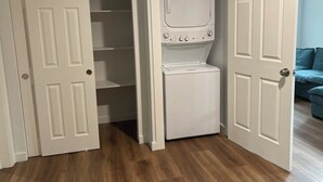 A laundry room with a washer and dryer, wooden flooring, and a blue couch in the adjacent room.