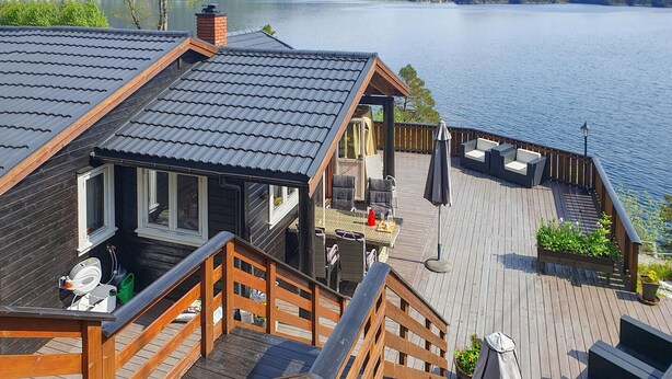 A wooden deck with a view of a lake, a covered outdoor seating area, and a staircase leading to the upper level. - Beach front home in Omastrand (Omastrand)