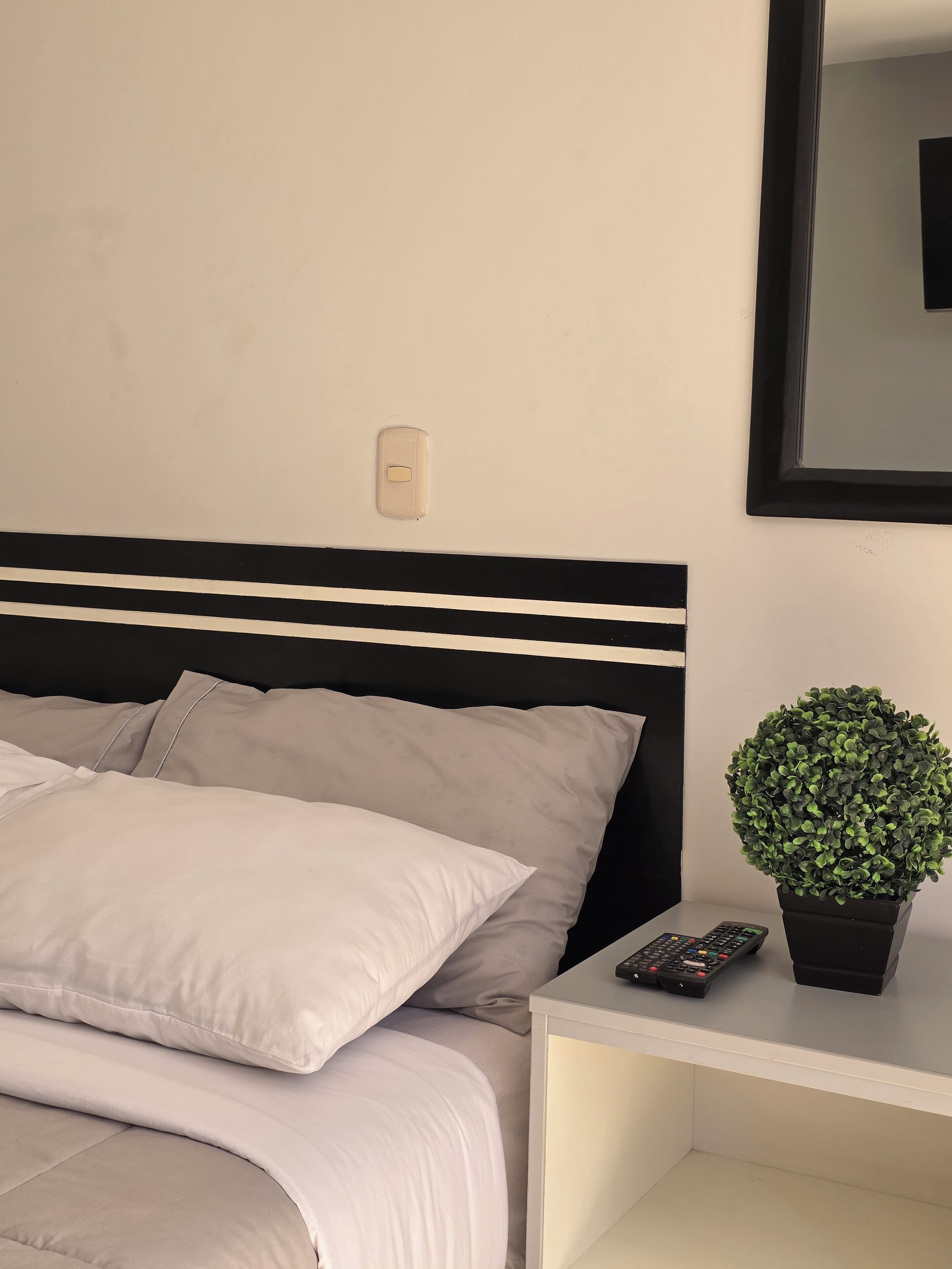 Una cama bien hecha con ropa de cama gris, un cabecero blanco y negro, una mesita de noche con un control remoto y una planta en maceta.