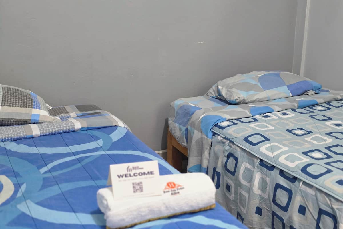 Zwei Einzelbetten mit blau gemusterten Bettdecken, ein Willkommensschild und ein zusammengerolltes Handtuch auf dem Bett links.