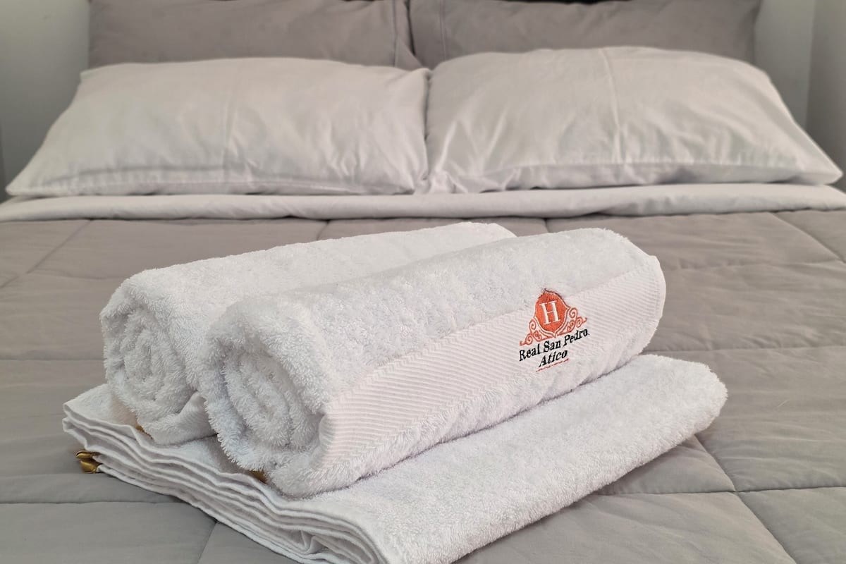 Ein ordentlich bezogenes Bett mit weißen Leinen und einem gefalteten Handtuch mit Logo.