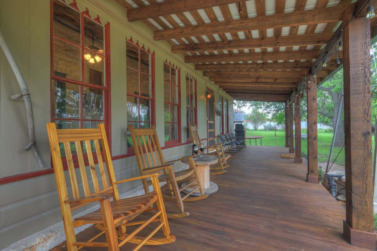 A covered outdoor seating area with wooden chairs and tables, wooden beams overhead, and large windows with red trim.