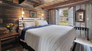 A rustic bedroom with a wooden headboard, a large bed with white bedding, a bedside table with a lamp, and a window with a view of the outdoors. - Rocking Ox Gasthaus and Barn Cabin (Fredericksburg)