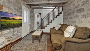A rustic living room with a stone wall, wooden ceiling, a brown sofa, a TV, and a staircase. - Rocking Ox Gasthaus and Barn Cabin (Fredericksburg)