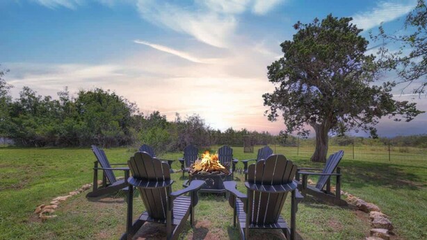 A fire pit with four Adirondack chairs arranged around it in a grassy outdoor area. - Rocking Ox Gasthaus and Barn Cabin (Fredericksburg)