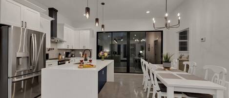 Una cocina moderna con una isla central, armarios blancos, electrodomésticos de acero inoxidable e iluminación colgante, además de un comedor con sillas blancas.