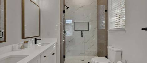 Et moderne badeværelse med et brusebad bag glas, et hvidt toilet og et stort spejl med lysarmatur over.