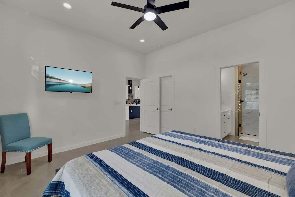 A bedroom with a bed, a ceiling fan, a TV, a chair, and a bathroom visible through an open door.