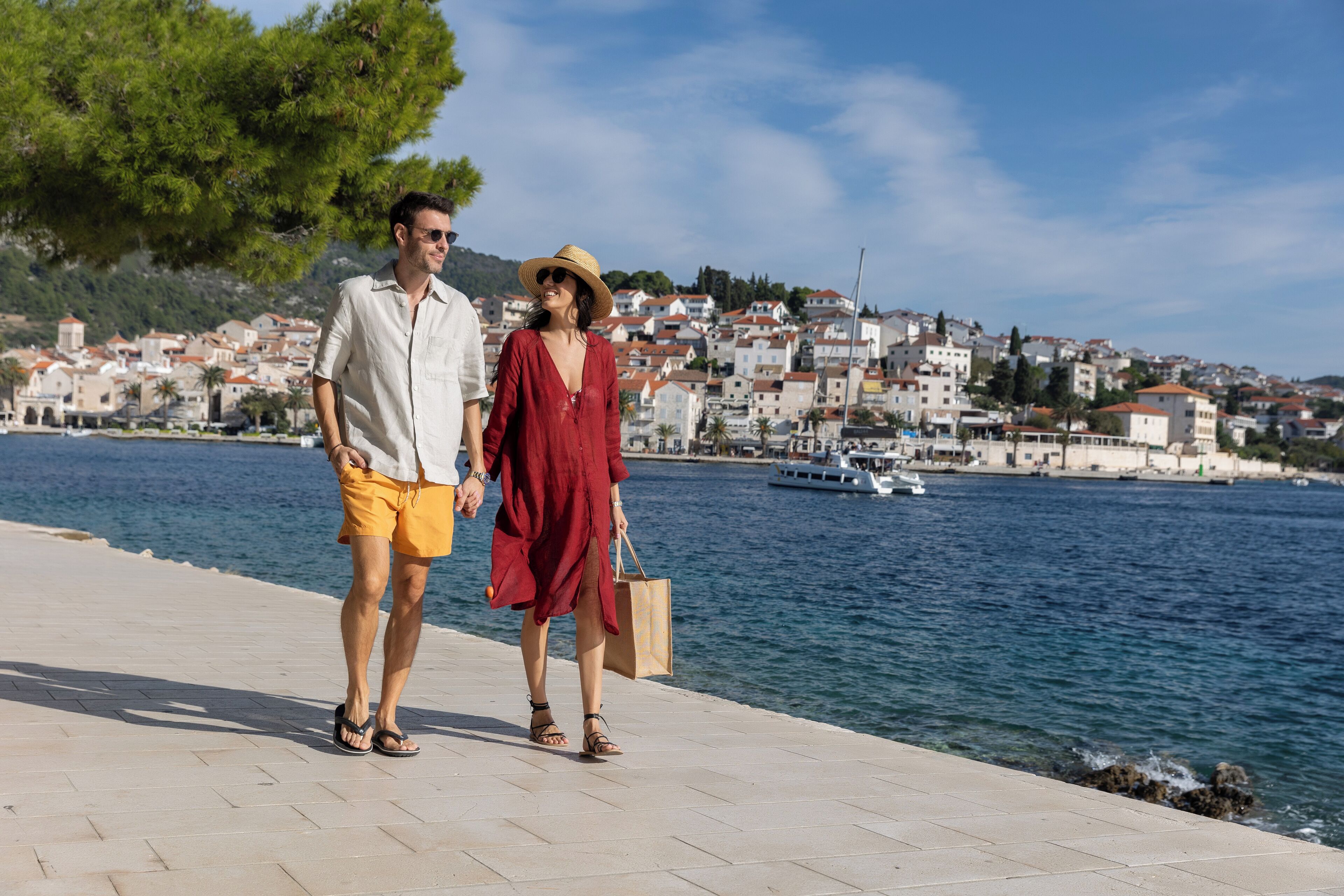 A couple walking along a waterfront promenade with a backdrop of coastal buildings and a boat in the water.