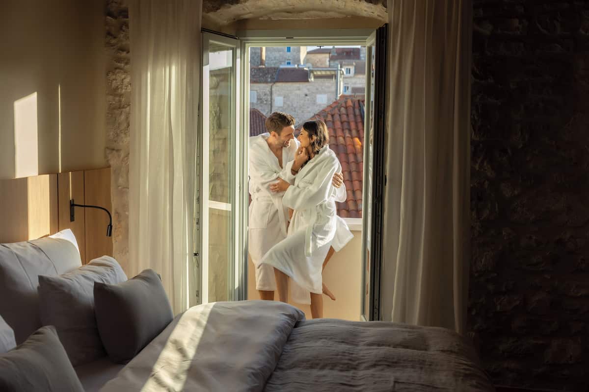 Una pareja vestida de blanco se abraza en una habitación con cama, pared de piedra y vistas a edificios a través de una puerta de cristal.
