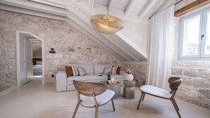 A modern loft with exposed stone walls, a high ceiling, and a large window. There is a white sofa, two wooden chairs, and a round coffee table with decorative items. - Forkira Luxury Hvar (Hvar)