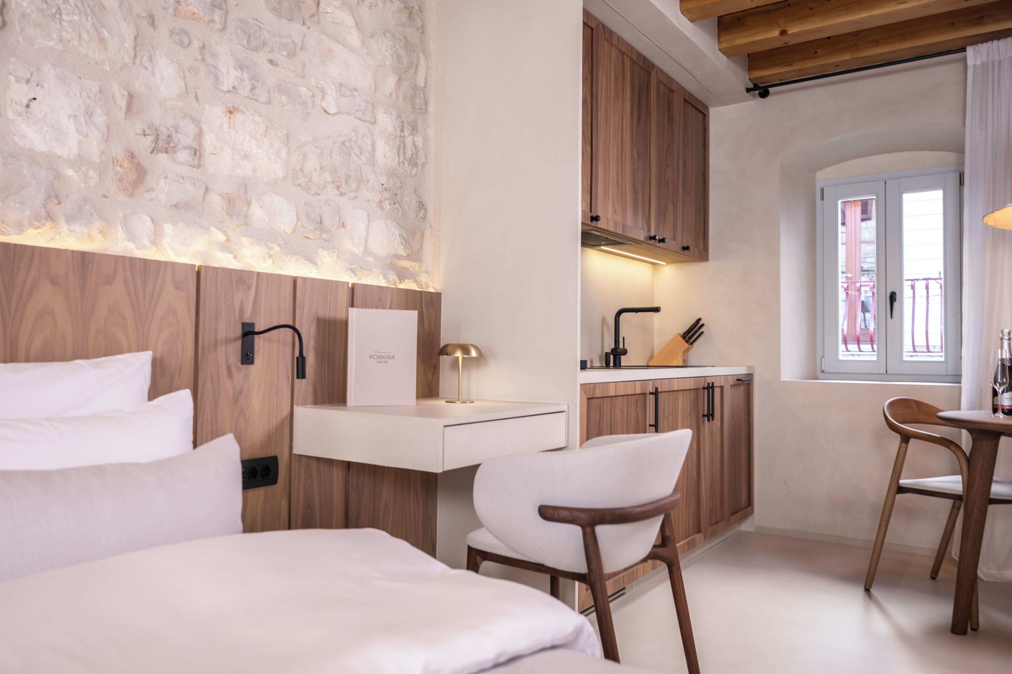 Une chambre d’hôtel moderne avec un mur en pierre, des meubles en bois, un lit blanc et une petite kitchenette.